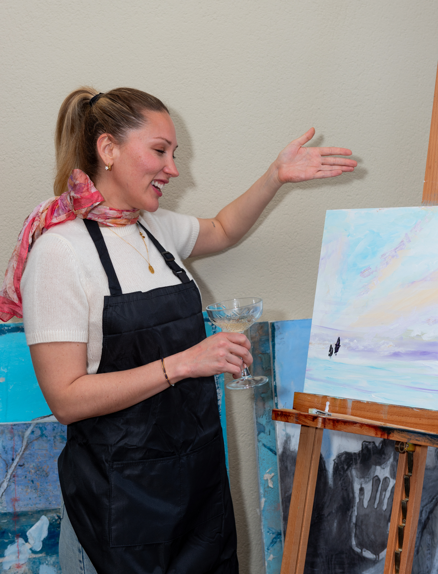 Smiling host in apron raising a coupe glass beside a fresh seascape painting